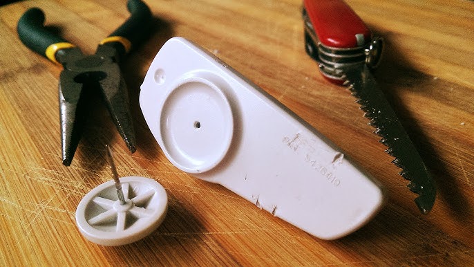 A disassembled ink tag lies on a wooden surface, with pliers and a multi-tool nearby, suggesting a DIY attempt to remove it.